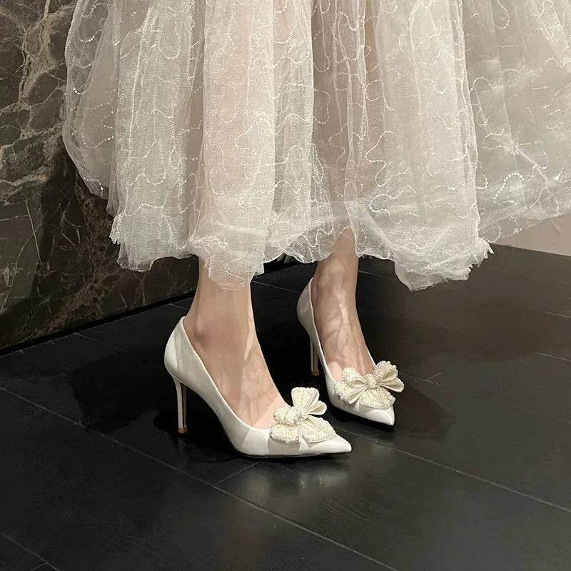 White satin stiletto heels with pointed toe and pearl bow detail, designed for brides by LYNHURD.